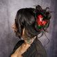 Woman with black hair and tattoos wearing a red floral hair accessory against a gray background