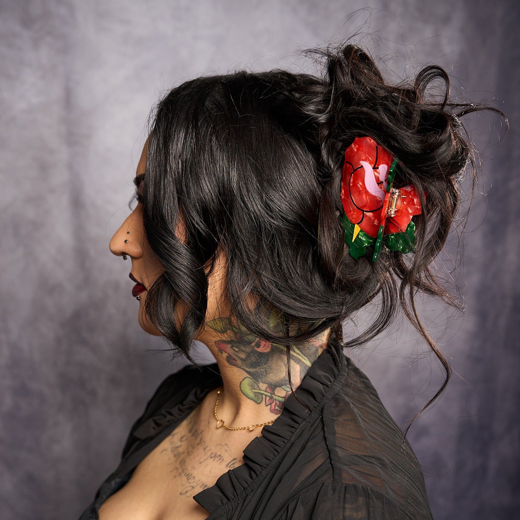 Woman with black hair and tattoos wearing a red floral hair accessory against a gray background